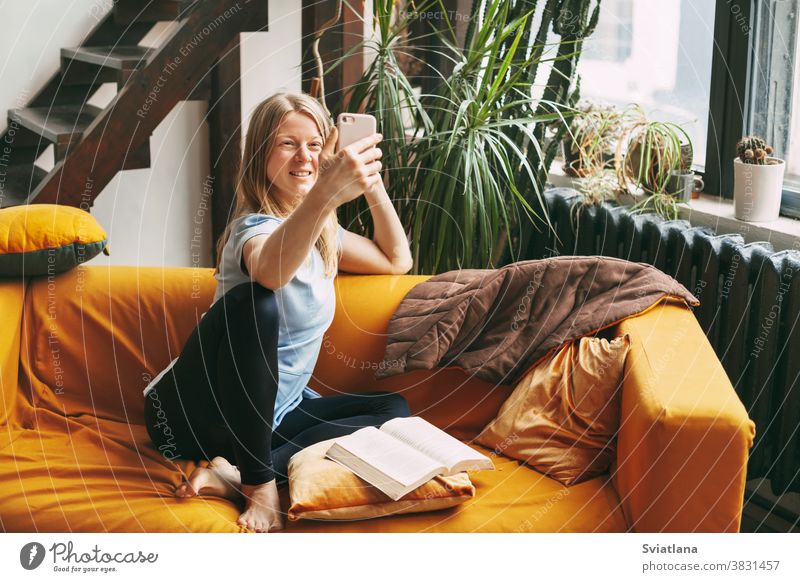 A young girl is sitting on the sofa and taking a selfie on her phone, an open book is lying next to her home woman cellphone room wireless text mobile internet