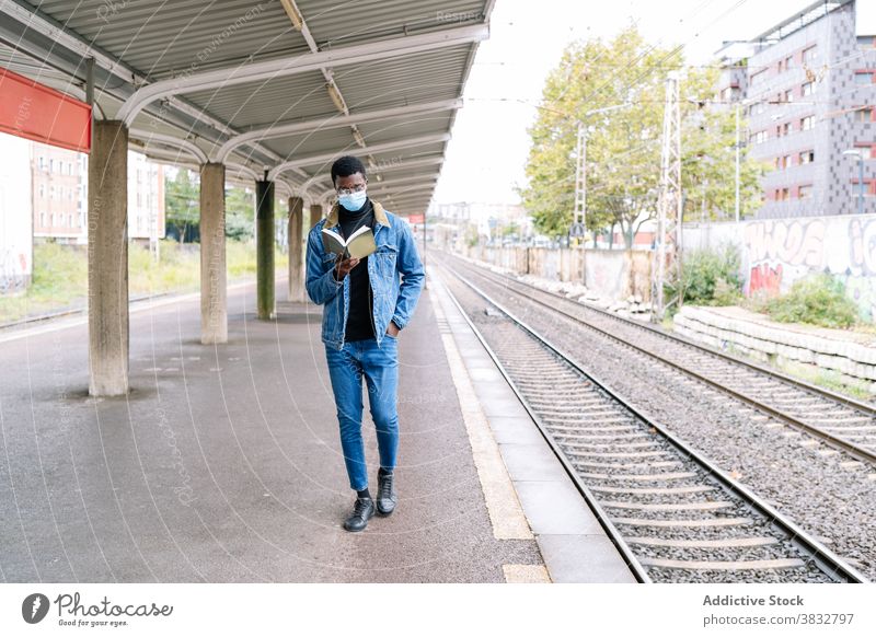 Black man with book on train platform wait railway new normal traveler coronavirus mask male ethnic black african american pandemic railroad transport station