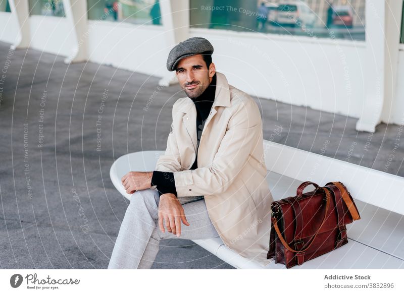 Serious man sitting on bench and looking at camera serious confident pensive style touch knee legs crossed building male thoughtful ethnic beard trendy bag