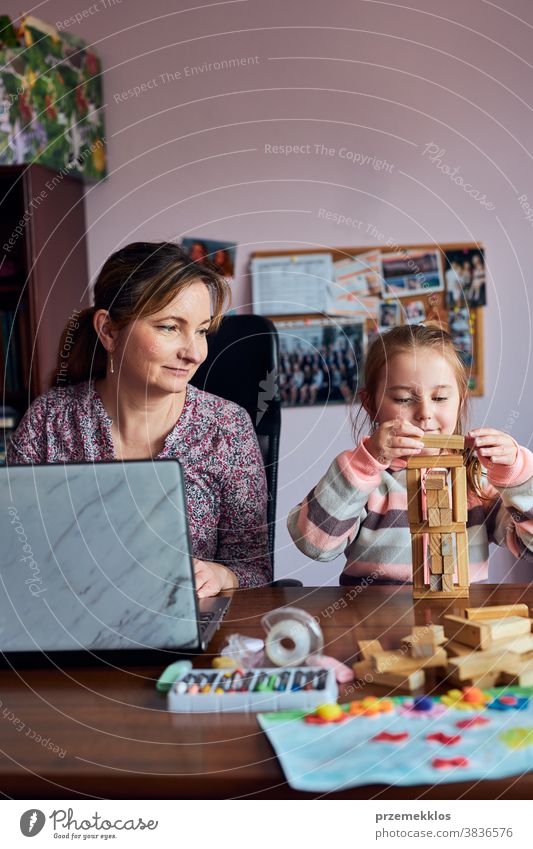 Woman mother working doing her job remotely during video chat call stream online course webinar on laptop from home while her daughter playing with bricks toy