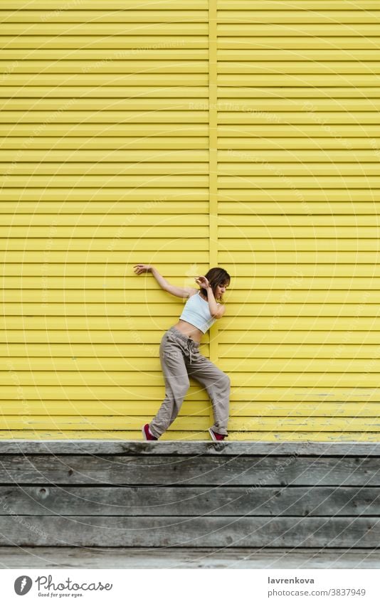 Young adult female in white top and grey pants dancing in front of bright yellow wall dancer choreographer hiphop outdoors girl stylish urban movement sport