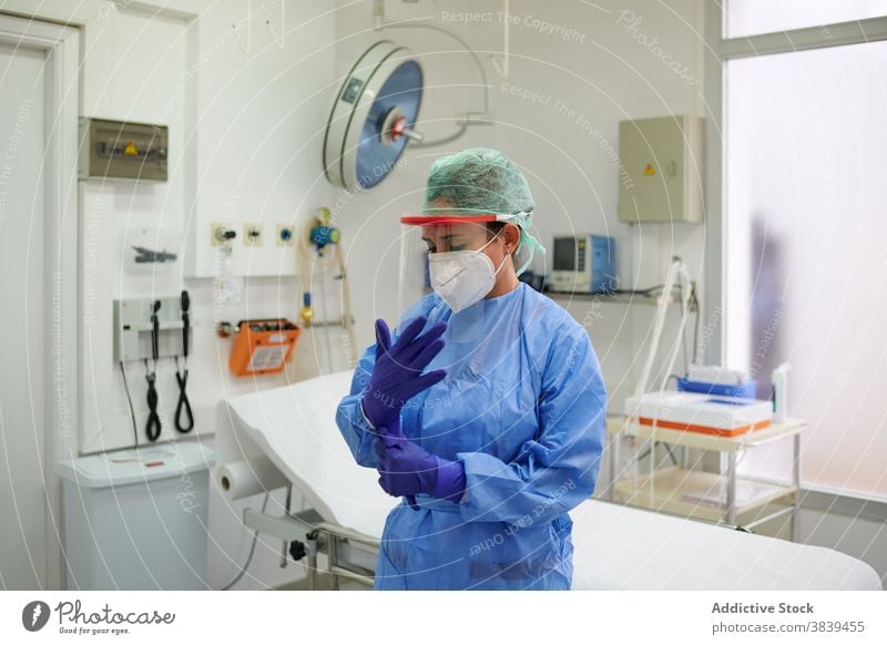 Anonymous doctor in uniform putting on sterile gloves put on job process equipment medical clinic woman mask respiratory hospital blue cap couch professional