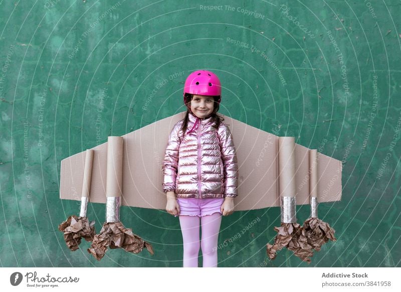 Smiling kid in cardboard jetpack looking at camera carton wing plane pilot child play achieve strength victory having fun celebrate success helmet cheerful girl