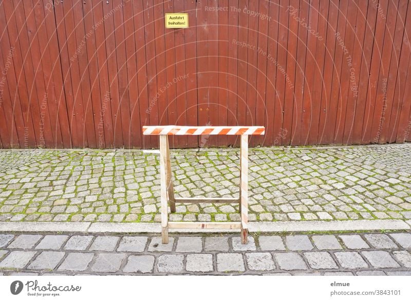 A wooden work trestle stands partly on the road and partly on the footpath in front of a dense, red-brown wooden fence with a yellow sign "Keep the exit free!