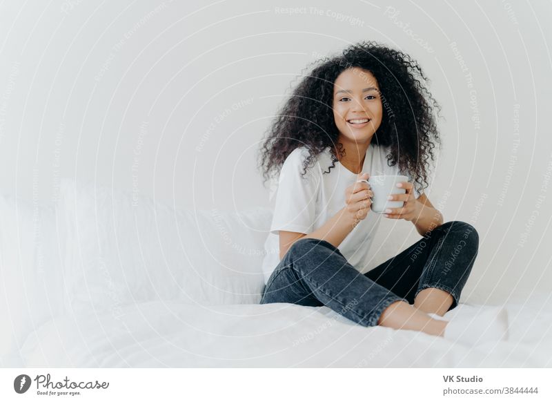 Young pretty African American woman rests in bed at home, drinks hot tea in morning, enjoys domestic atmosphere, tasty drink while relaxes in bedroom. People, leisure, rest and lifestyle concept