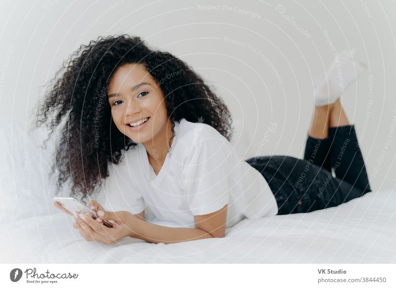 Horizontal shot of curly ethnic woman with pleasant smile, lies on comfortable bed, raises legs, uses modern cellphone, reads text message, enjoys high speed internet connection. People and bed time