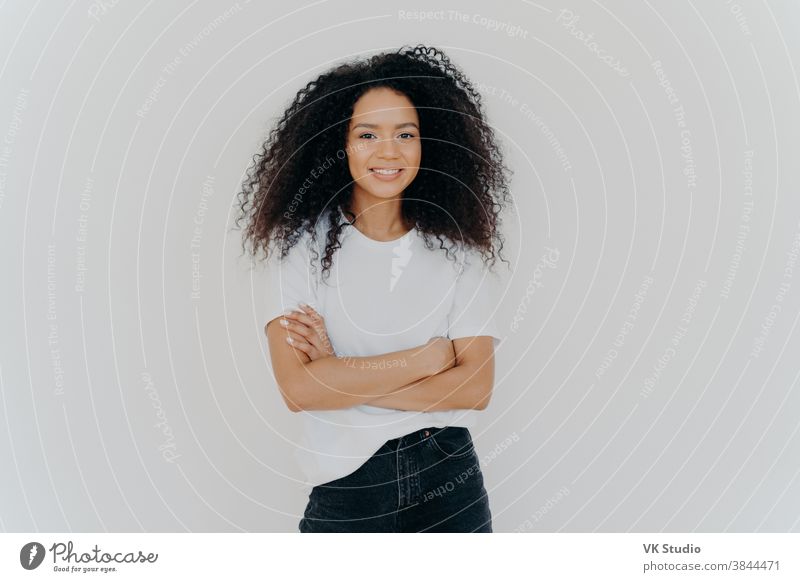 Studio shot of carefree young beautiful woman with Afro hairstyle, keeps arms folded, smiles joyfully, wears casual t shirt and jeans, isolated on white background. People, ethnicity, face expressions