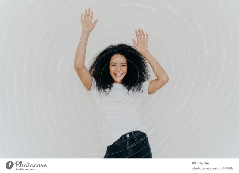 Half length shot of energetic carefree curly woman cheers with hands raised, wears white t shirt and jeans, has lucky day, perfect weekend, models against white background. Everybody put your hands up