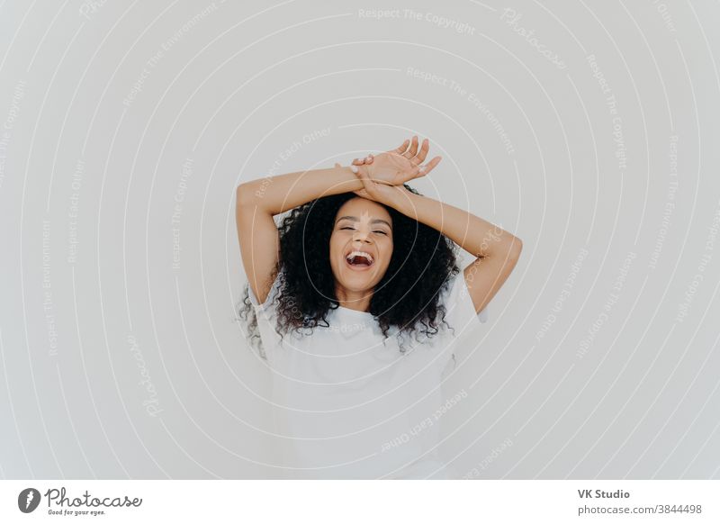 Overjoyed curly haired young lady keeps both hands on forehead, opens mouth and laughs happily, feels energetic, dressed in casual wear, poses against white studio background. What funny joke!