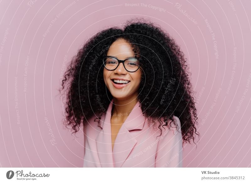 Positive curly haired businesswoman wears spectacles, formal wear, has bushy hairstyle, being in good mood after successful day at work, poses against violet background. Monochrome. Happy director