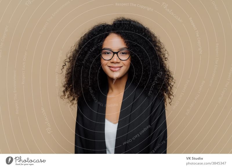 Portrait of beautiful Afro American woman with crisp hair, dressed in elegant black jacket, transparent glasses, looks directly at camera with gentle smile wears optical glasses isolated on brown wall