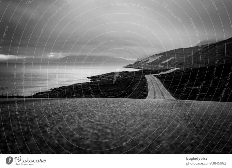 Lonely winding coastal road on which a car is only visible in the distance. To the left is the fjord/the sea and to the right and in the background mountains. The sky is cloudy and dull.