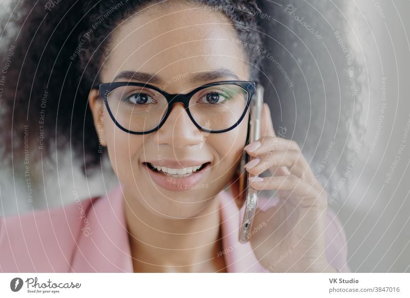 Close up portrait of happy dark skinned businesswoman holds mobile phone near ear, calls business partner, has charming smile, wears optical glasses, has curly bushy hair, discusses working issues