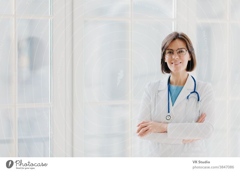 Self confident professional doctor stands with arms crossed, wears white medical gown with stethoscope, thinks about work positively, poses against big window. Healthcare and occupation concept