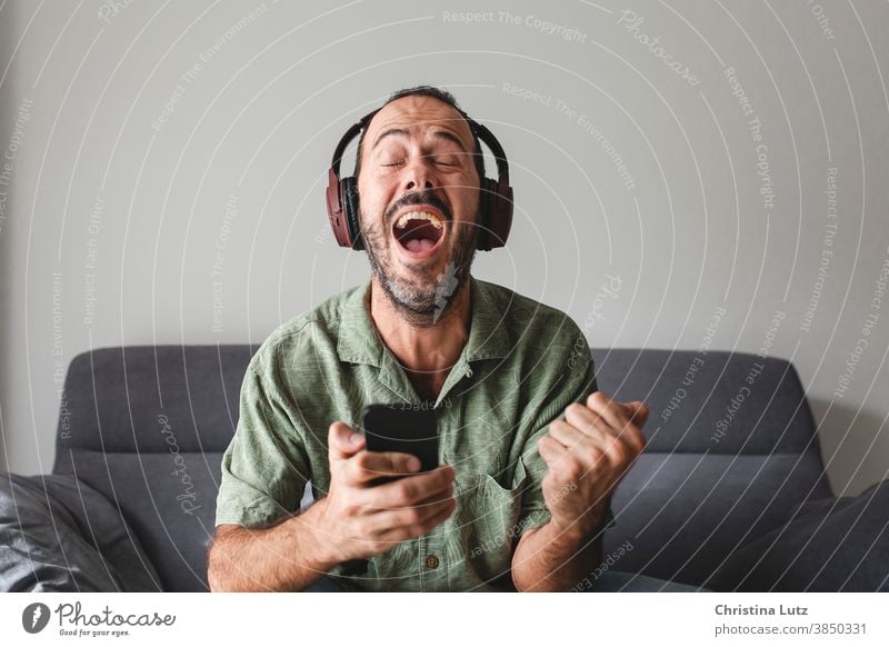 man sitting on sofa with mobile phone and headphones, shouting for joy while winning sports betting scream for joy smartphone results enthusiastic closed eyes