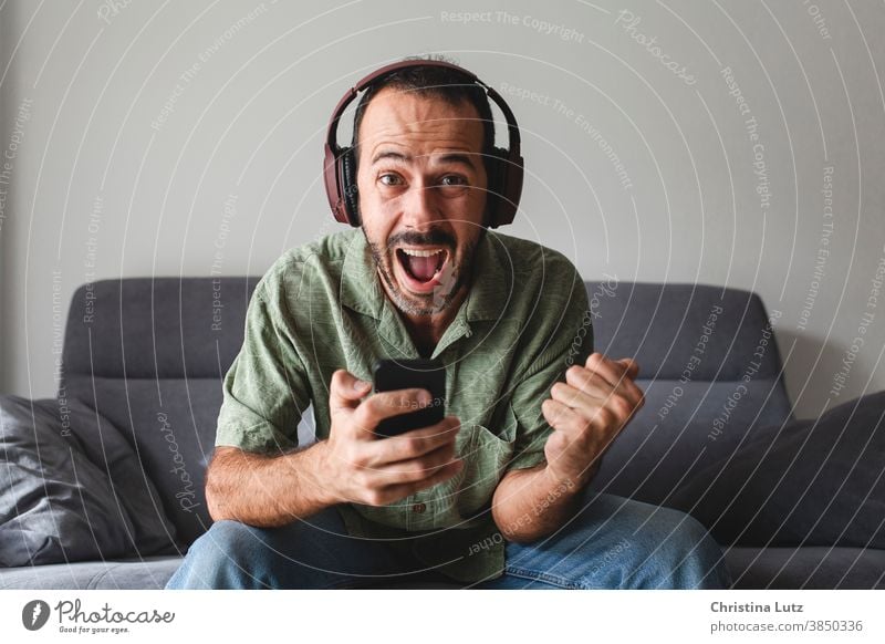 man sitting on sofa and shouting for joy, while winning sports betting with mobile phone scream for joy clenched fist yelling headphones enthusiastic results