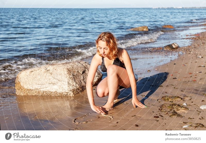 young girl in a swimsuit draws with her finger on the sand by the seashore beauty happy beautiful drawing sunny female water coast ocean holiday wave summer