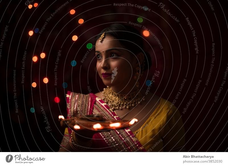 An young and beautiful Indian Bengali woman in Indian traditional dress is holding a Diwali diya/lamp in her hand in front of colorful bokeh lights. Indian lifestyle and Diwali celebration