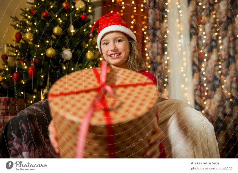 A happy child in red Santa hat is celebrating Christmas. Cute girl holding wrapped gift, smiling and waving hand, sitting in decorated with Christmas lights and tree room. Happy holidays