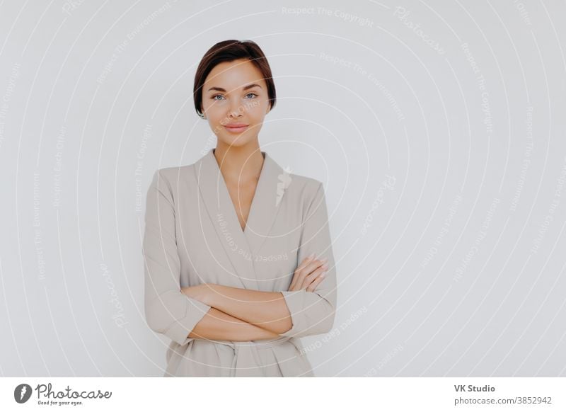 Prosperous successful business lady wears elegant wear, crosses hands while stands indoor at office over white wall, looks directly at camera. Female entrepreneur models indoor, waits for partner