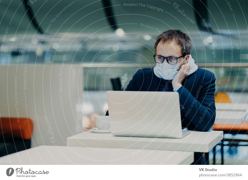 Photo of serious man concentrated in screen of laptop computer, works from distance during coronavirus outbreak, wears protective mask not to spread disease, drinks coffee, sits at white table