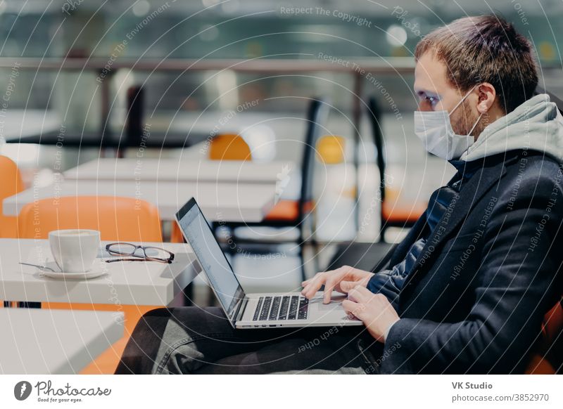 Indoor shot of male freelancer works freelance on modern laptop computer, wears medical mask against coronavirus, poses in cafe, concentrated at screen, reads news, connected to wireless internet