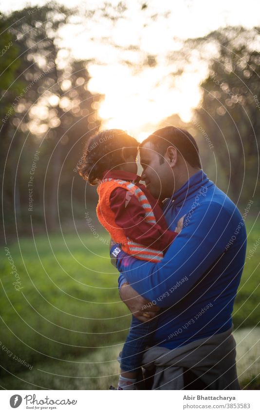 An Indian Telugu brunette father and his baby boy in winter garments enjoying themselves in winter afternoon on a  green grass field in forest background. Indian lifestyle and back light photography.