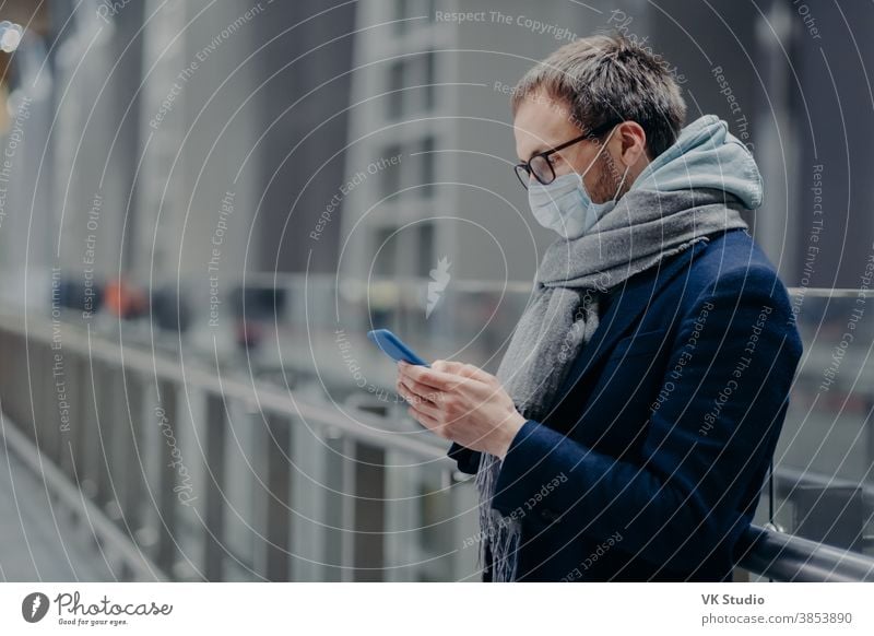 Sideways shot of male blogger sends text messages and reads comments from followers on modern cellphone, wears sterile medical mask to protect himself from coronavirus or other infectious disease