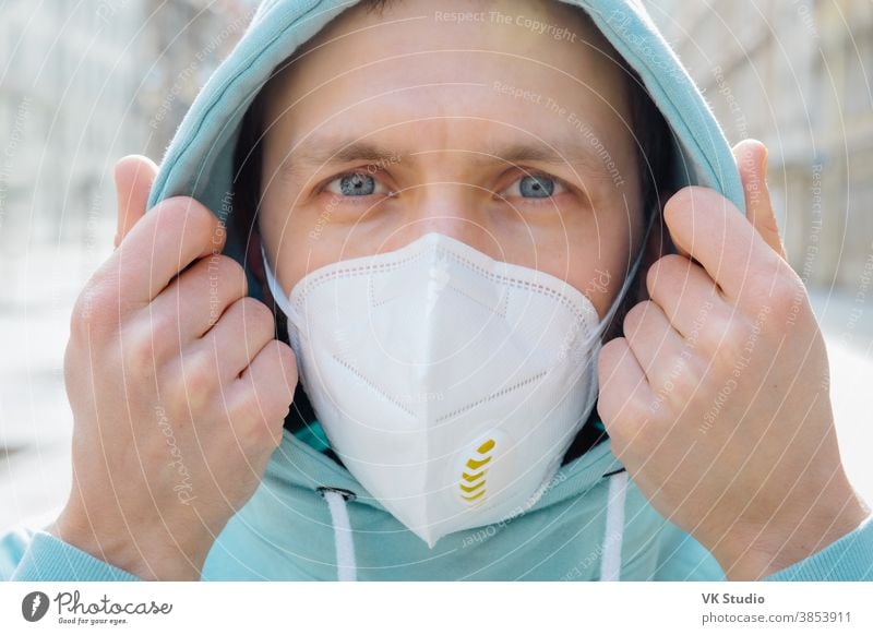 Close up shot of European man protects face with respiratory mask, wears hoodie, poses outdoor on street, looks seriosuly at camera, prevents coronavirus or Covid-19, flu. Street during quarantine