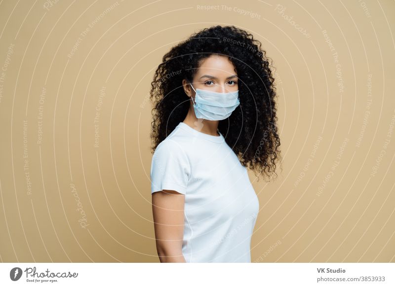 Sideways shot of Afro American woman wears protective face mask, protects against spread of coronavirus disease, stays at home, follows rules of self isolation. Covid-19, safety, quarantine concept