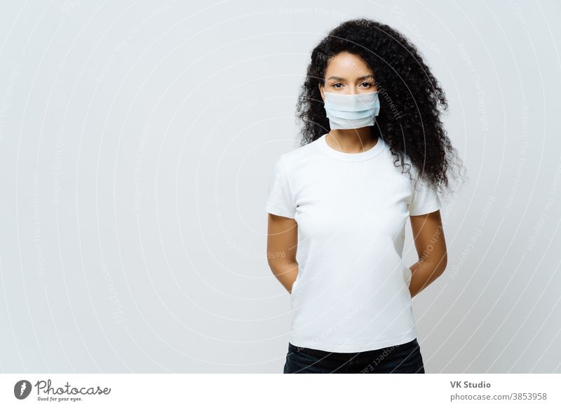 Half length shot of Afro American woman being on self isolation or quarantine, wears medical mask during coronavirus outbreak, listens news, tries not to spread virus, stays at home for long period