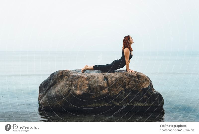 Young woman practicing yoga, at sea, in rain. Redhead girl meditating on a rock in the Baltic Sea, Germany 20s Iyengar yoga adult alone baltic sea caucasian