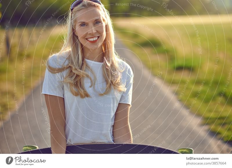 Portrait of a fit beautiful middle-aged woman with an active lifestyle smiling and looking at camera while holding a longboard on a sunny road in the park in summer