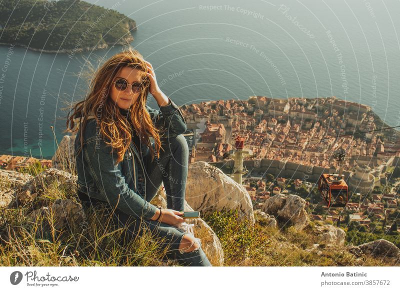 Attractive brunette sitting on the Srd hill above the city of Dubrovnik, observing the city in the distance. Wind blowing her hair above the ancient famous landmark town