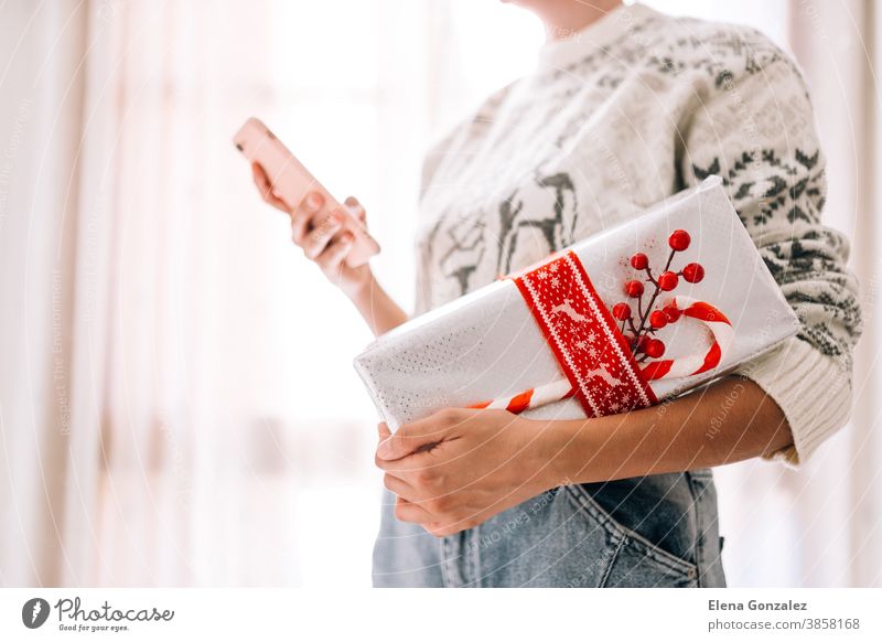 Young unrecognizable woman watching her mobile phone holds in a gift box in metallic paper with red twine and candy. Christmas New Years present. christmas