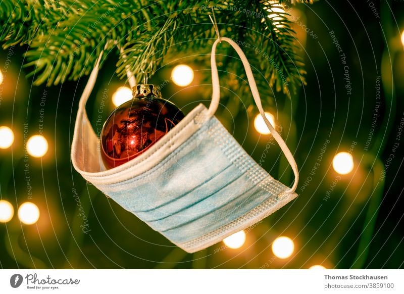 Red Christmas tree ball and a face mask on a branch from the Christmas tree, lights in the background. Symbol of the impact of the Covid 19 virus in the Christmas season 2020 and 2021.