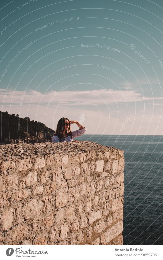 Vertical shot of a brunette standing on the city walls of the Dubrovnik town, holding her hand over her eyes and looking into the distance as if she is searching for something