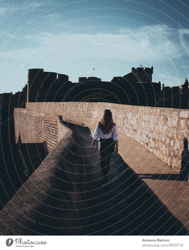 Vertical shot and rear view of an attractive girl walking along the city walls of Dubrovnik town. Dark moody edit with black and orange highlights holiday relax