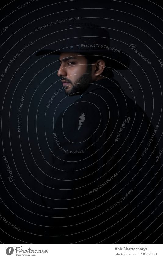 Fashion portrait of young and handsome Indian Bengali brunette man with striped formal shirt, black suit and hat turning back in black copy space background. Indian lifestyle and fashion photography.