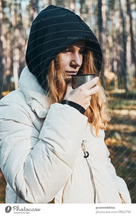 Woman in a hood having break during autumn trip holding cup with hot drink from thermos flask on autumn cold day active activity adventure coffee destination