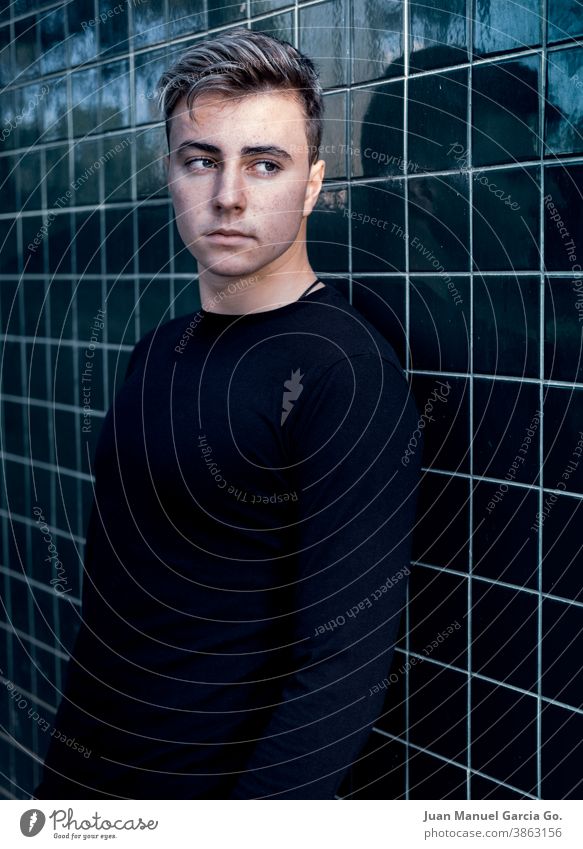 Young teenager with light-colored hair, wearing a black shirt leans against a square tiled wall latin american and hispanic ethnicity 14-15 years hairstyle