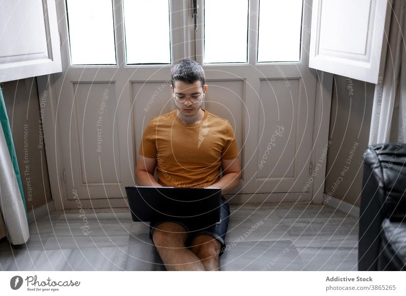 Busy man working on laptop in living room freelance remote home typing project concentrate browsing male casual outfit floor sit window legs crossed device job