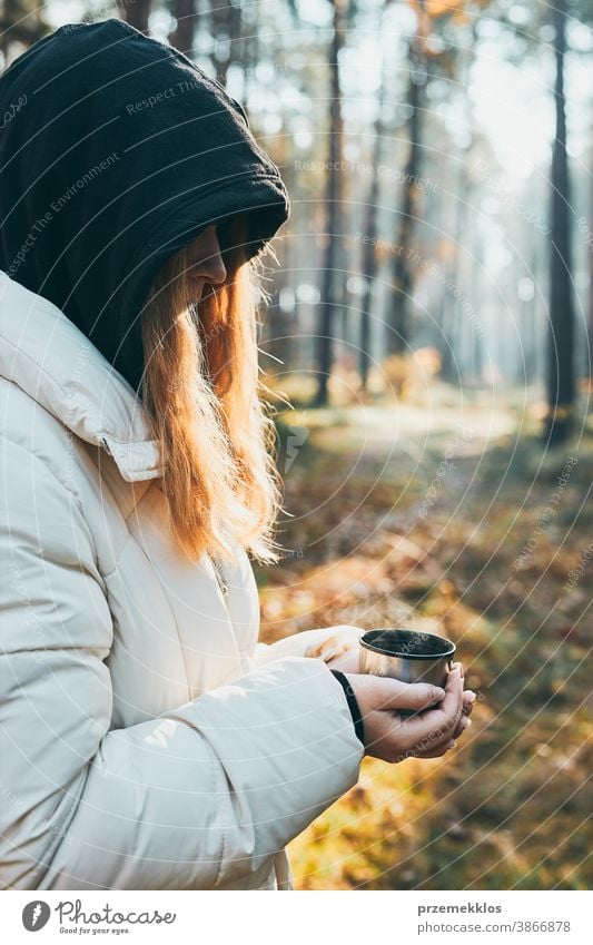 Woman in a hood having break during autumn trip holding cup with hot drink from thermos flask on autumn cold day. Active girl wandering in a forest actively spending time