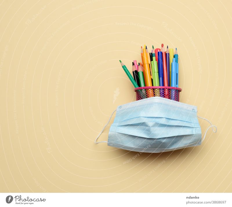 disposable medical mask and multi-colored pencils and pens in a metal bucket on a beige background pandemic prevention protective quarantine safe safety school