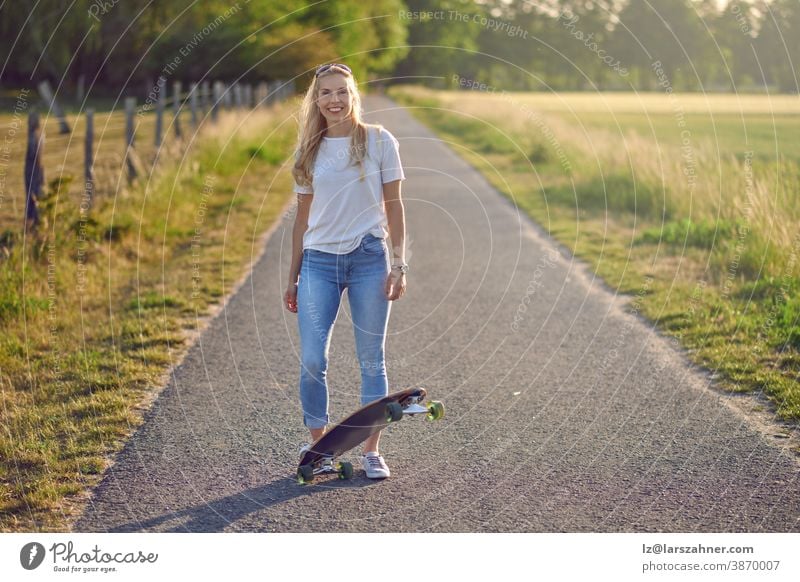 Attractive athletic blond woman with her skateboard backlit by the evening sun on a rural road between fields smiling at the camera middle-aged fit playing