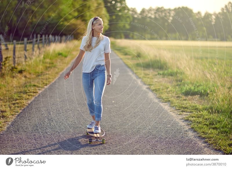 Fit active middle-aged woman playing on her skateboard approaching the camera along a narrow rural road with a happy smile backlit by the evening sun fit blond