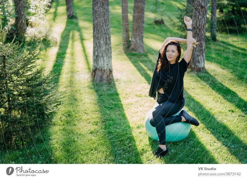 Photo of active flexible brunette woman stretches with fitball, does pilates or fitness exercises, poses on green grass during sunny spring day in forest, enjoys gymnastic exercises on fresh air.