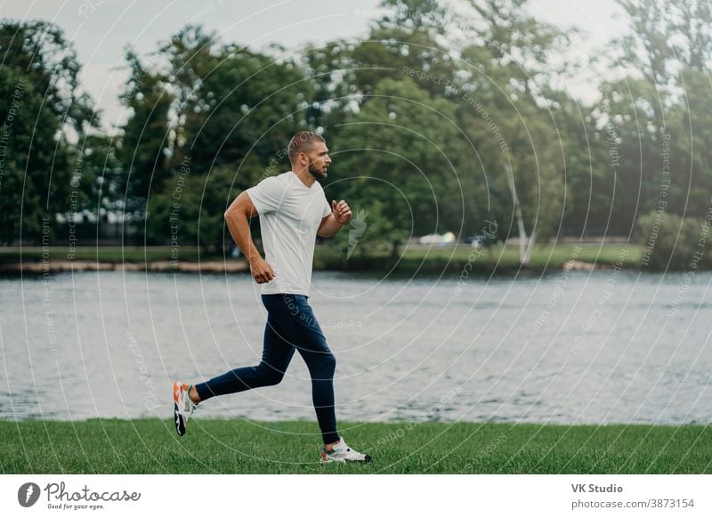 Athlete bearded man runs along beautiful landscape, demonstrates endurance, dressed in active wear, goes jogging near river, breathes fresh air. Sport, exercising, fitness and workout concept