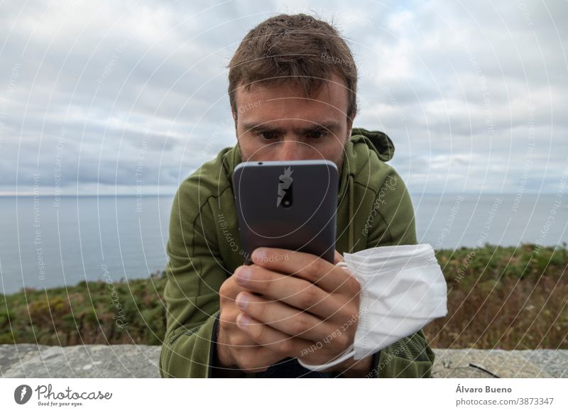 A young man, grabbing a face mask and a smart mobile phone, Spain. protective new normal chat looking internet digital telecommunications call gray hands