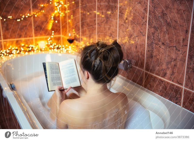 Young beautiful woman reading a book and drinking red wine in the bathtub with bubble foam, decorated with colorful lights, relaxing and spa concept young